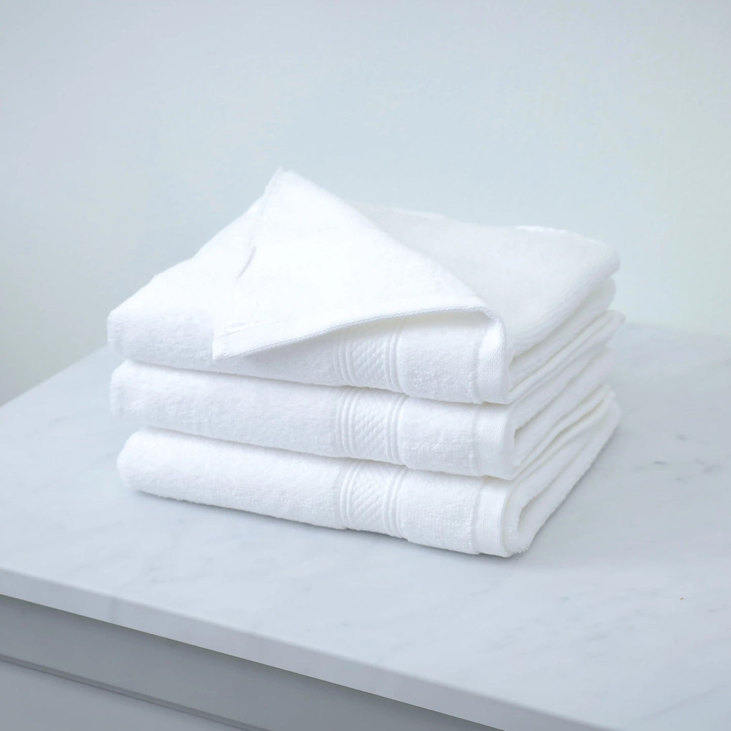 Valiant Products Atelier Zen white cotton bath towels with elegant dobby border, stacked on a marble surface.