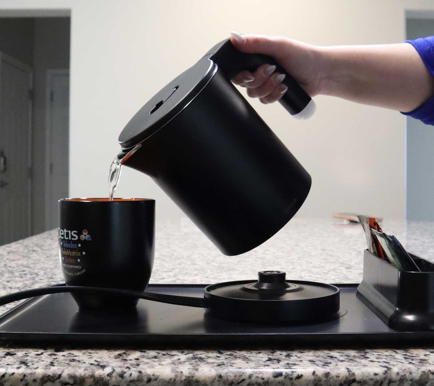 Valiant Products K-608 black electric kettle pouring hot water into a mug on a hotel hospitality tray