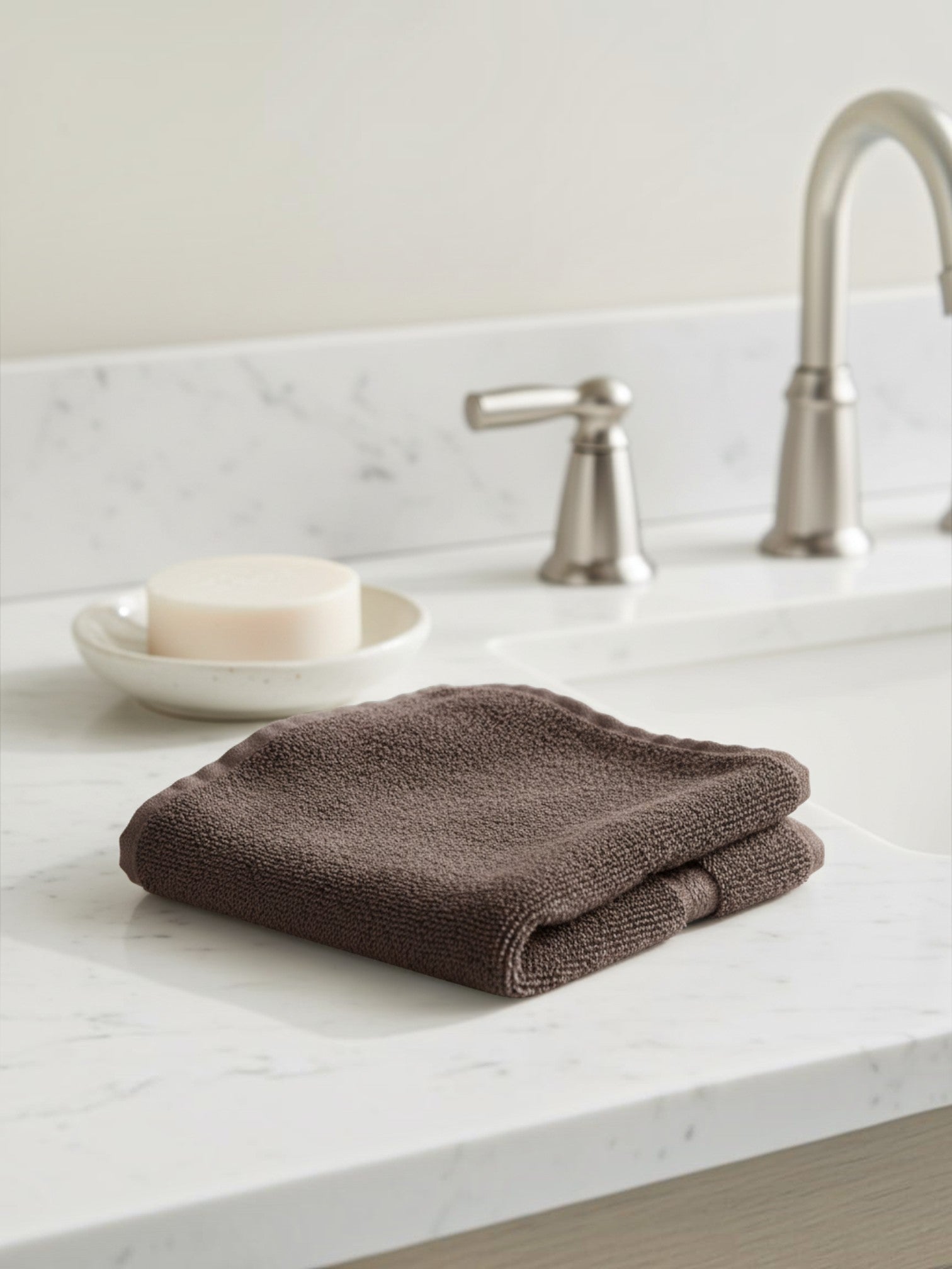 Valiant Products brown ring-spun cotton washcloth with a dobby border, resting on a white marble bathroom vanity.