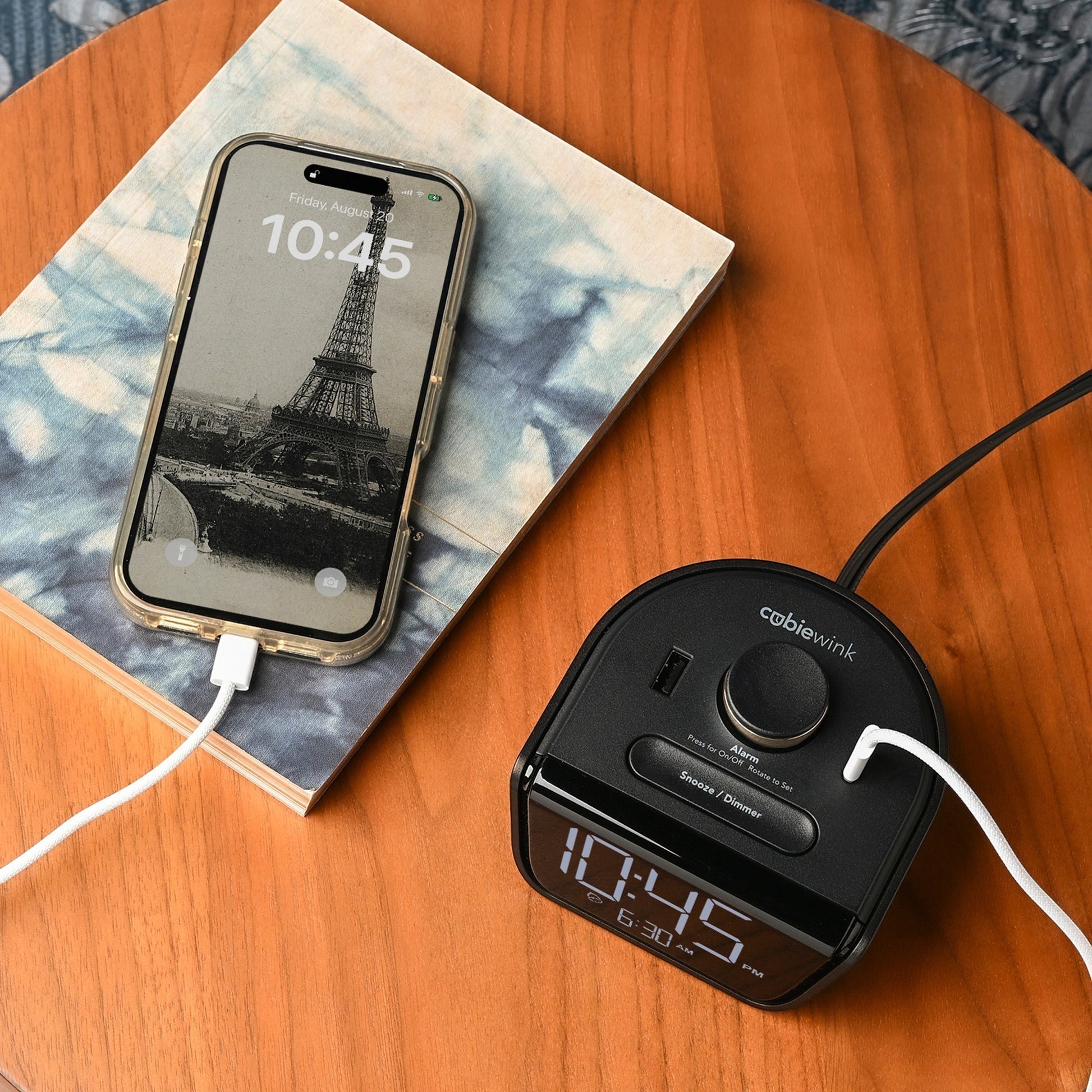 Valiant Products CubieWink black alarm clock with USB-A and USB-C ports charging a smartphone on a wooden nightstand
