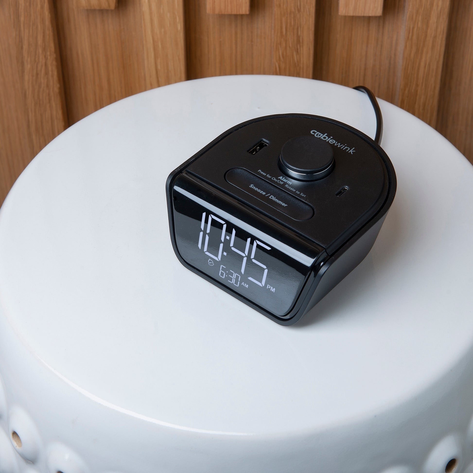Valiant Products CubieWink black compact hotel alarm clock with USB-A and USB-C charging ports on a white stool