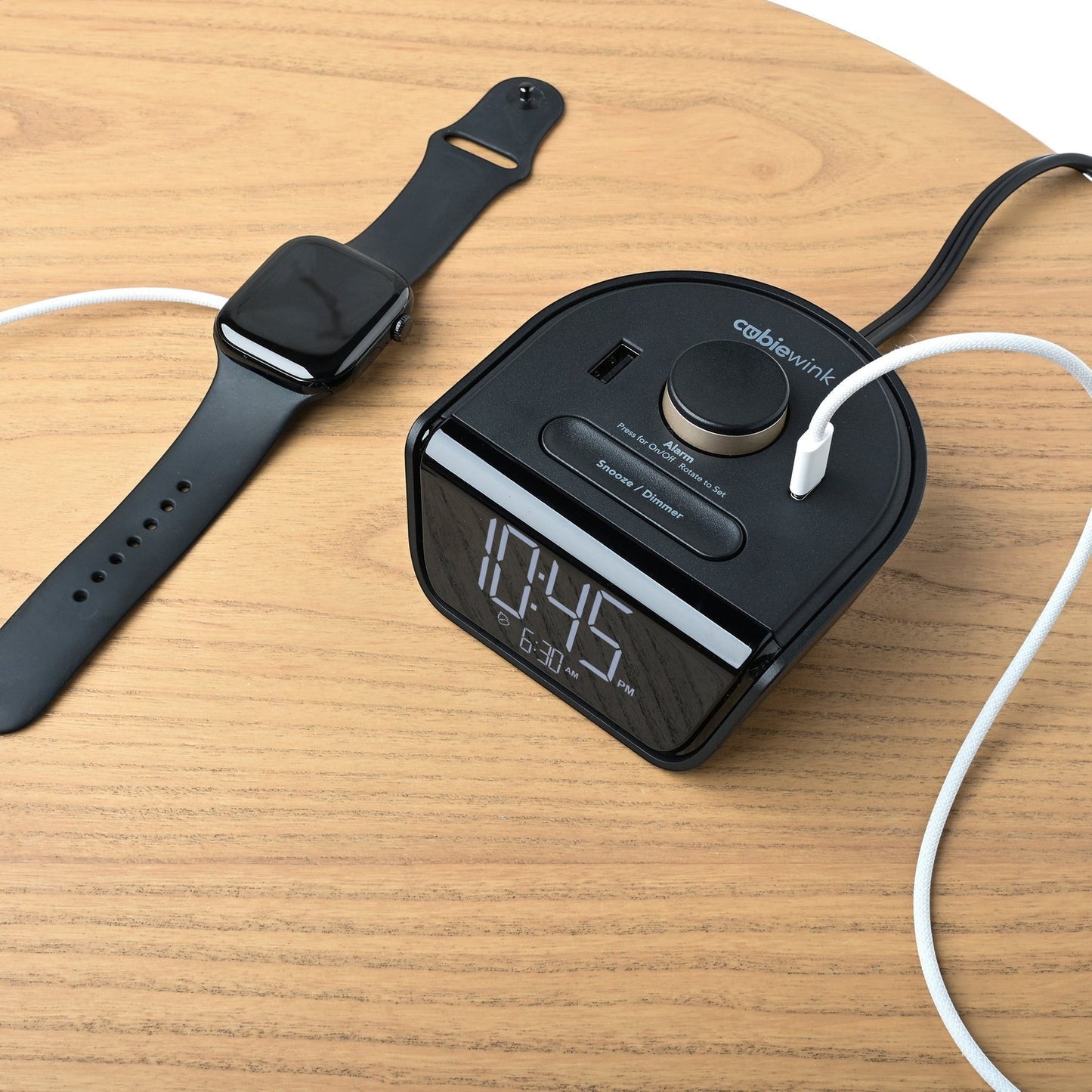 Valiant Products CubieWink black alarm clock with USB-A and USB-C ports charging a smartwatch on a wooden table.
