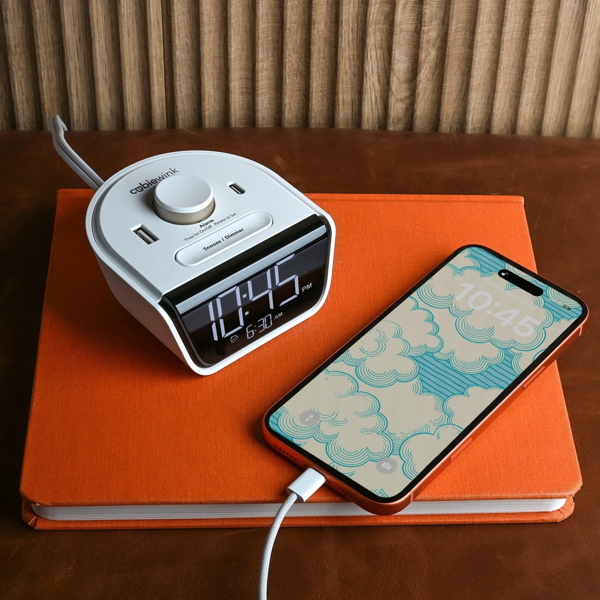 Valiant Products CubieWink white alarm clock with USB-C and USB-A ports charging a smartphone on an orange notebook.