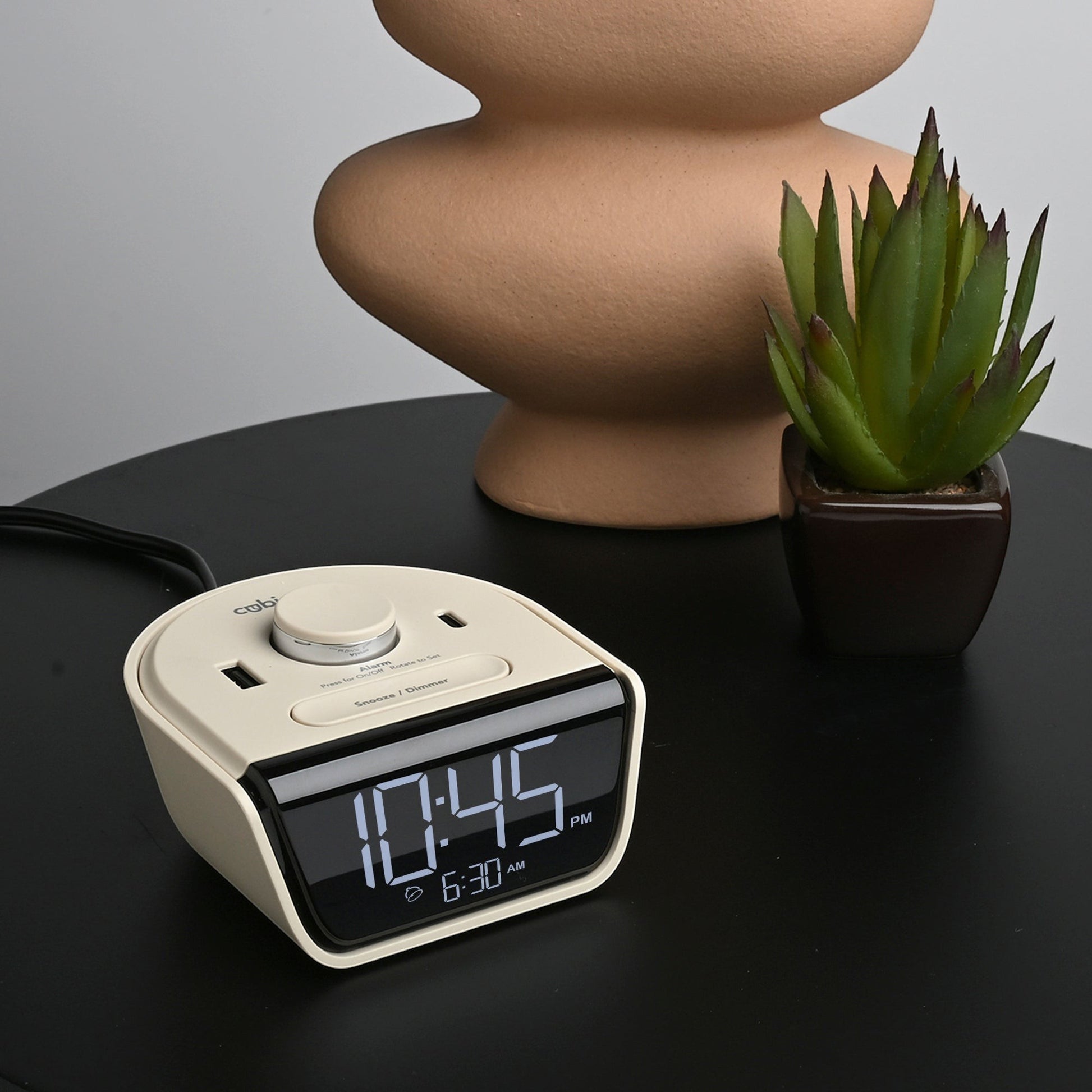 Valiant Products CubieWink latte-colored compact alarm clock with USB-A and USB-C ports on a black nightstand.