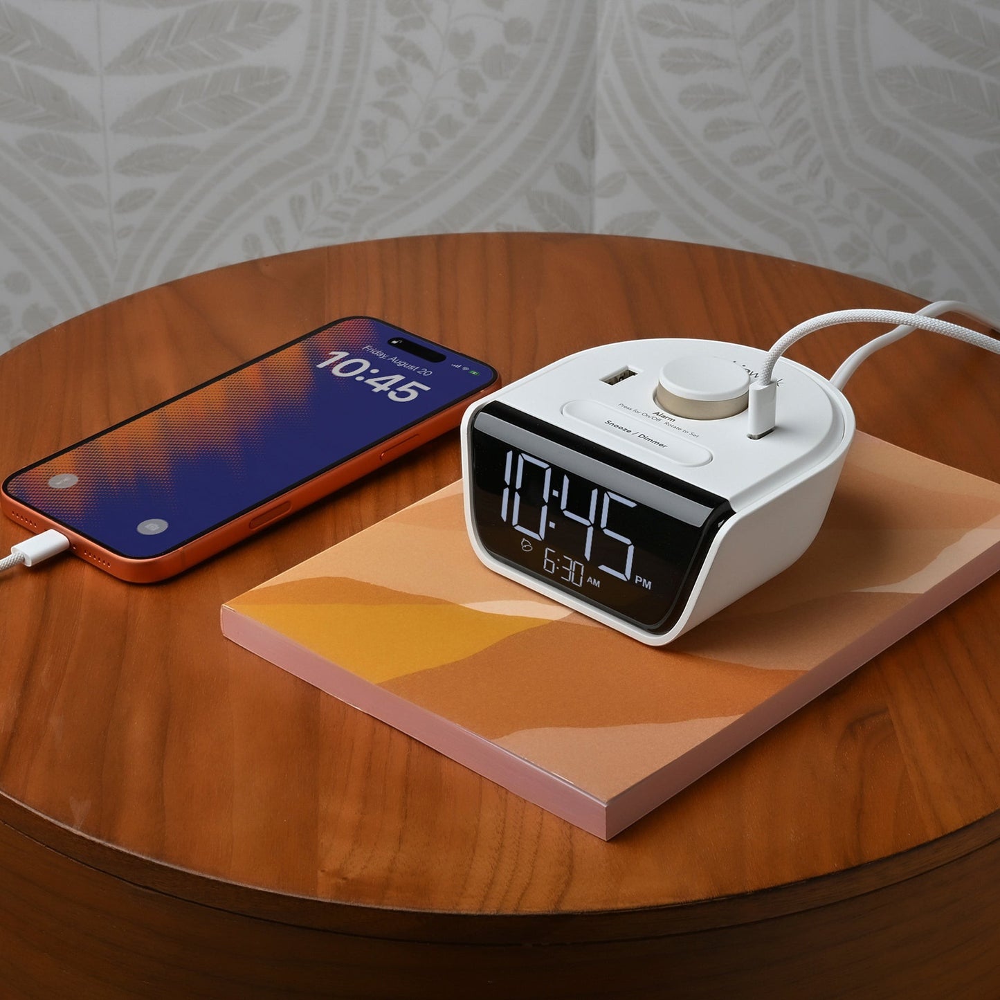 Valiant Products CubieWink white alarm clock with USB-A and USB-C ports charging a smartphone on a wooden nightstand.