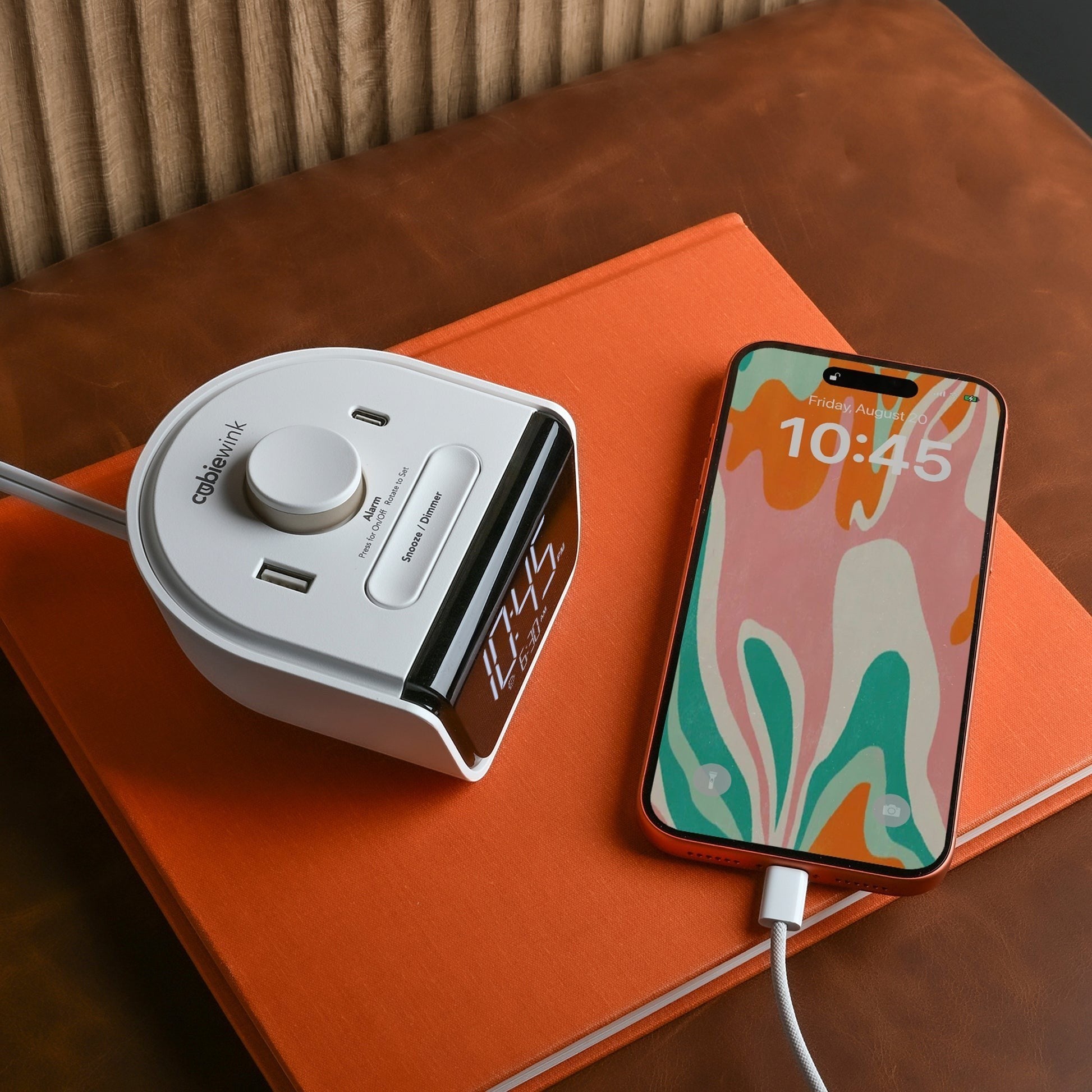 Valiant Products CubieWink white compact alarm clock with USB-C and USB-A ports charging a smartphone on a book.