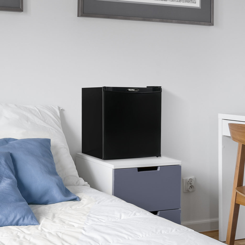 Valiant Products Danby 1.7 cu. ft. compact black refrigerator sitting on a white nightstand in a bedroom.