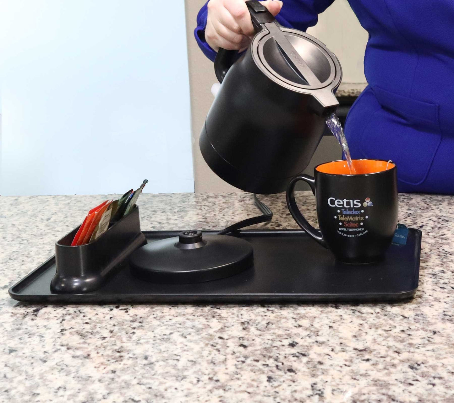 Valiant Products K-100 black electric kettle pouring hot water into a mug on a hospitality tray