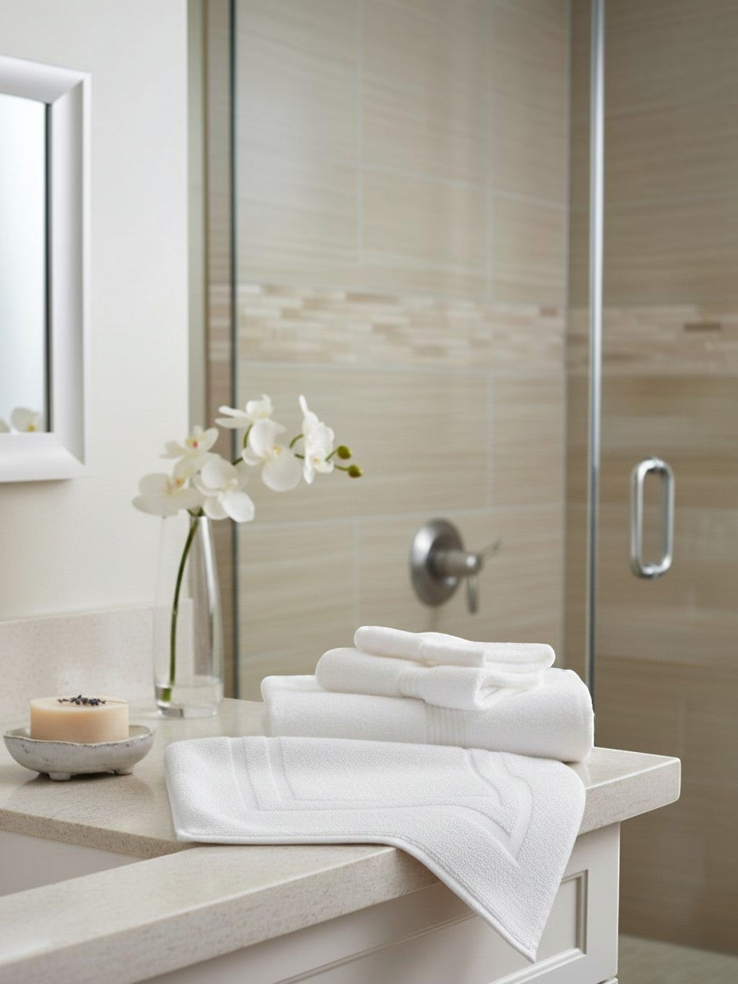 Valiant Products Sovereign white dobby border bath mats stacked on a bathroom vanity with orchid decor