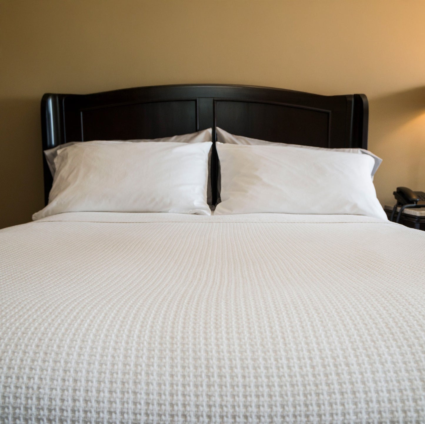 Valiant Products white cotton basketweave blanket on a made bed with a dark wood headboard in a hotel room setting.