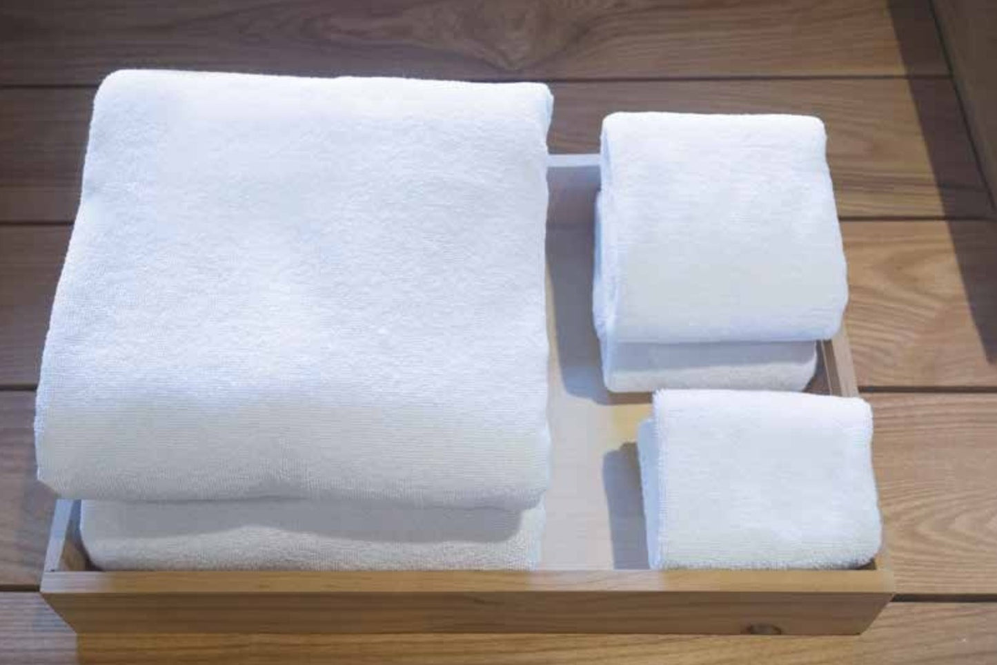 Valiant Products white Royal Suite Dobby border bath towels folded neatly in a wooden tray on a wooden surface